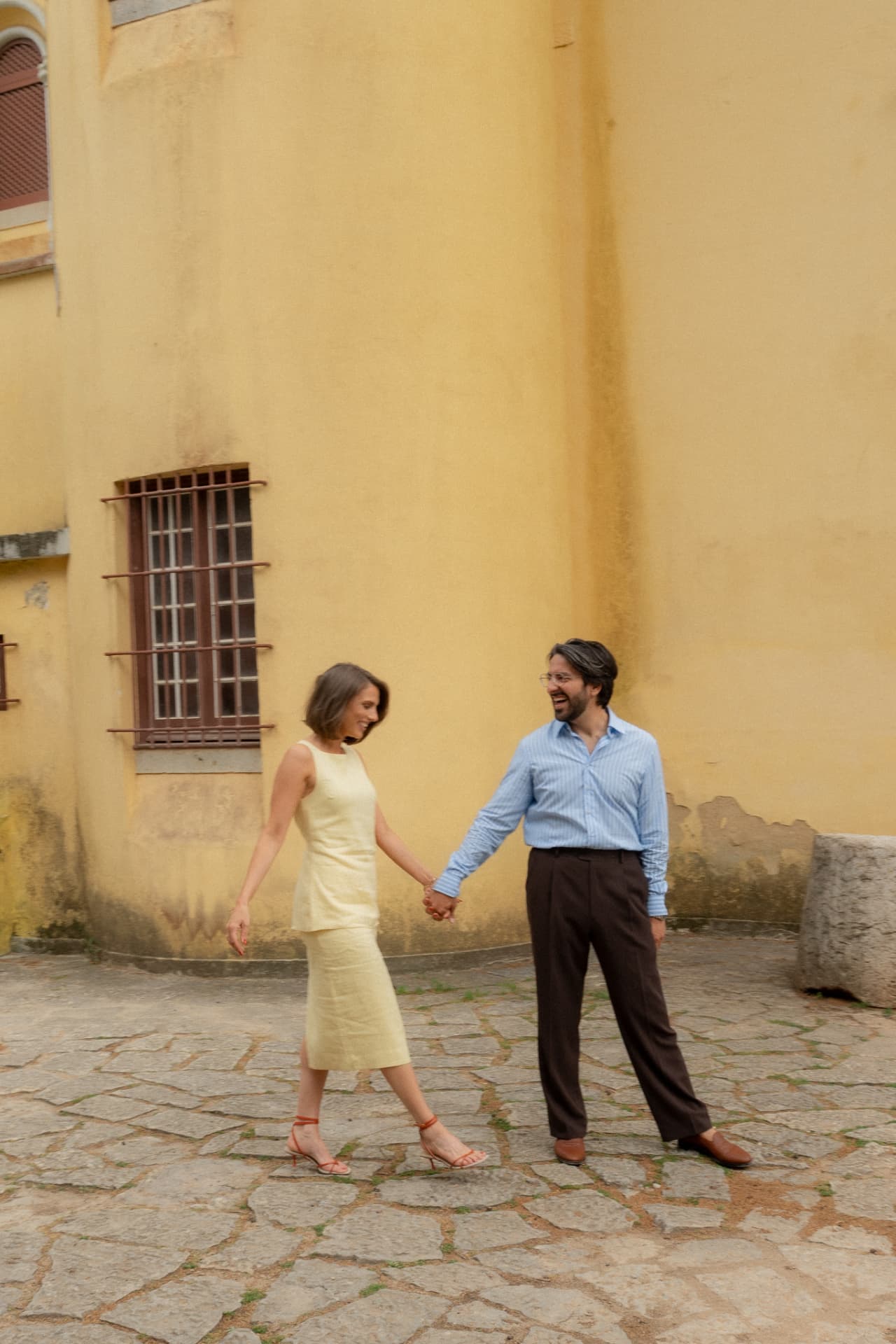 Holding hands on cobblestones beside a sunlit ochre wall